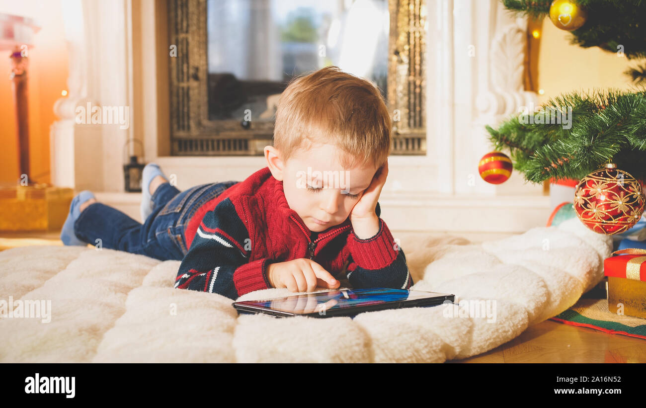 Getönten Closeup Portrait von kleinen Jungen liegen unter Weihnachtsbaum auf einem weichen Teppich und Spielen auf digital Tablet-PC Stockfoto