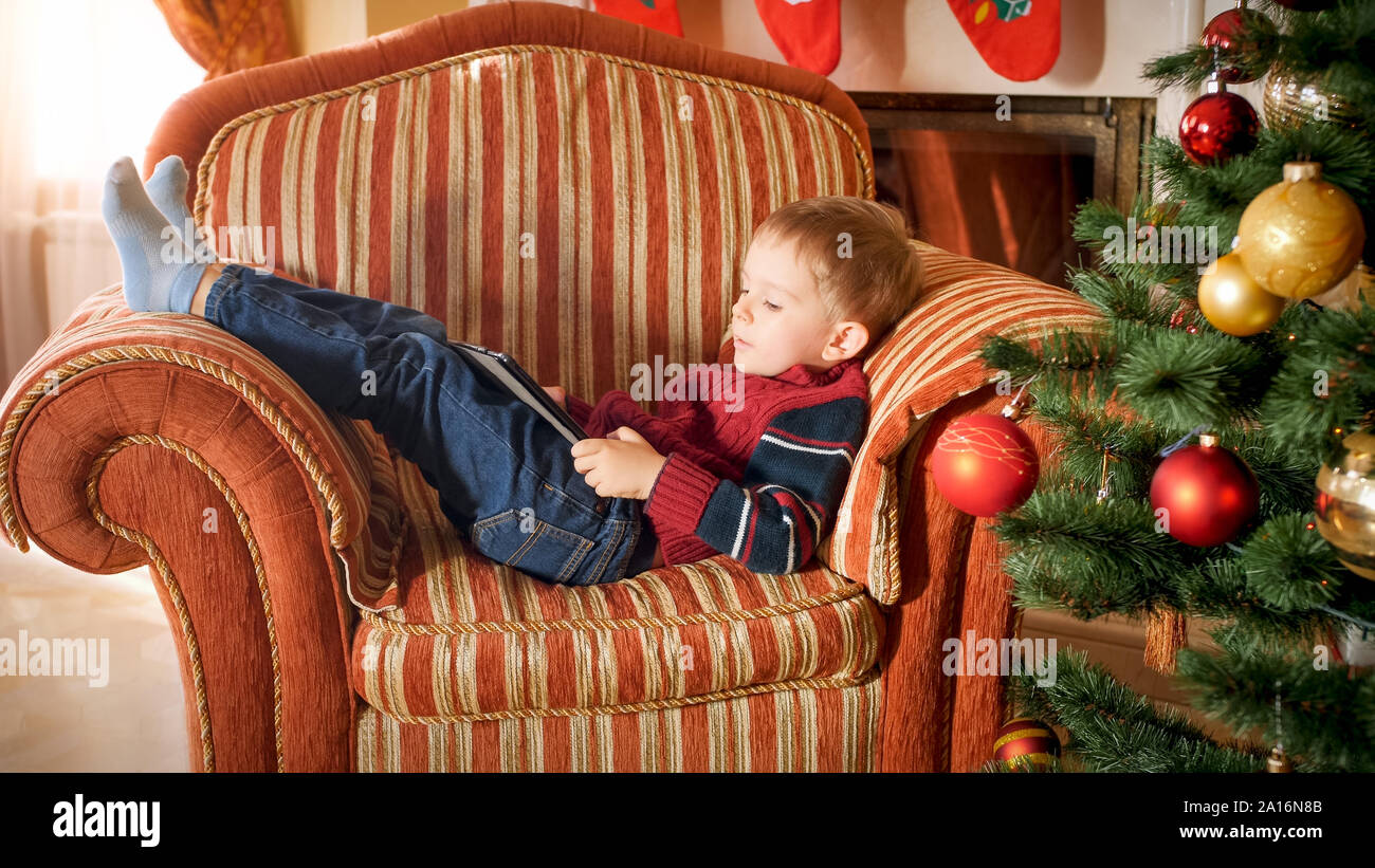 Portrait von kleinen Jungen im Sessel liegend neben schönen geschmückten Weihnachtsbaum und Spielen auf digital Tablet-PC Stockfoto