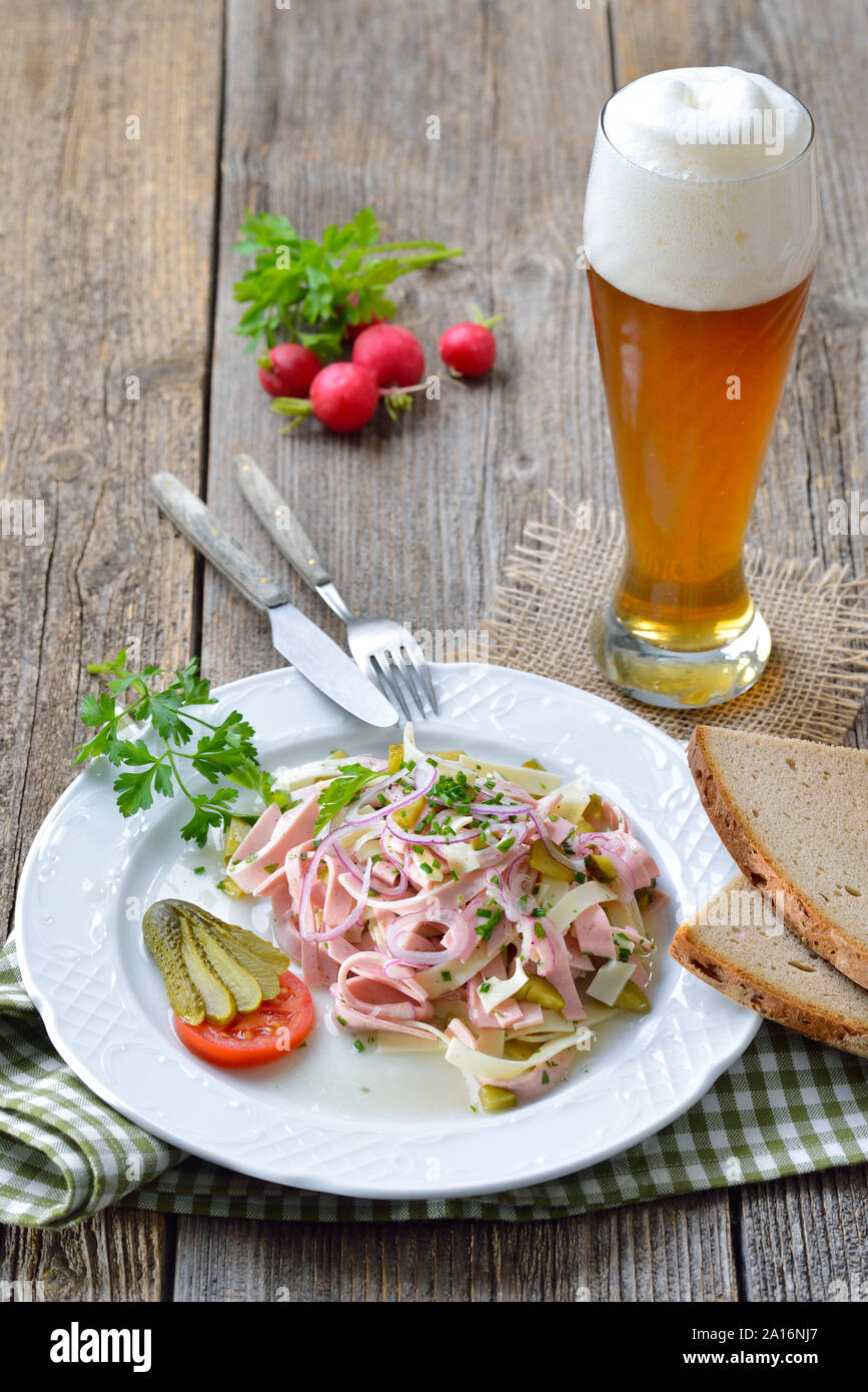 Herzhafte bayerische Wurstsalat mit Käse Streifen, Essiggurken und rustikales Bauernbrot serviert mit einem kühlen Weißbier Stockfoto