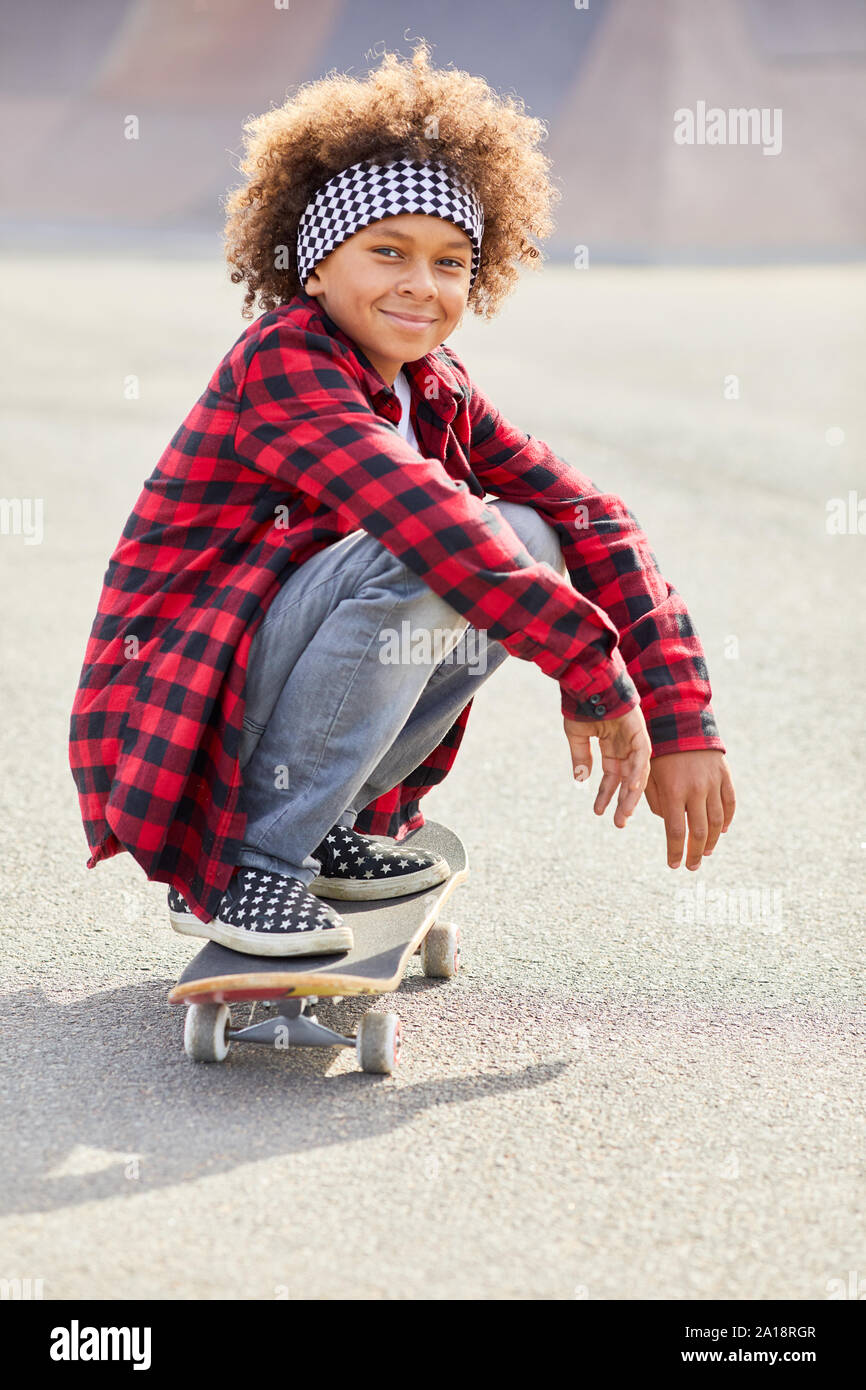 Portrait von afrikanischen Jungen mit dem lockigen Haar und in Freizeitkleidung Lächeln auf die Kamera beim Reiten auf Skateboard Natur Stockfoto