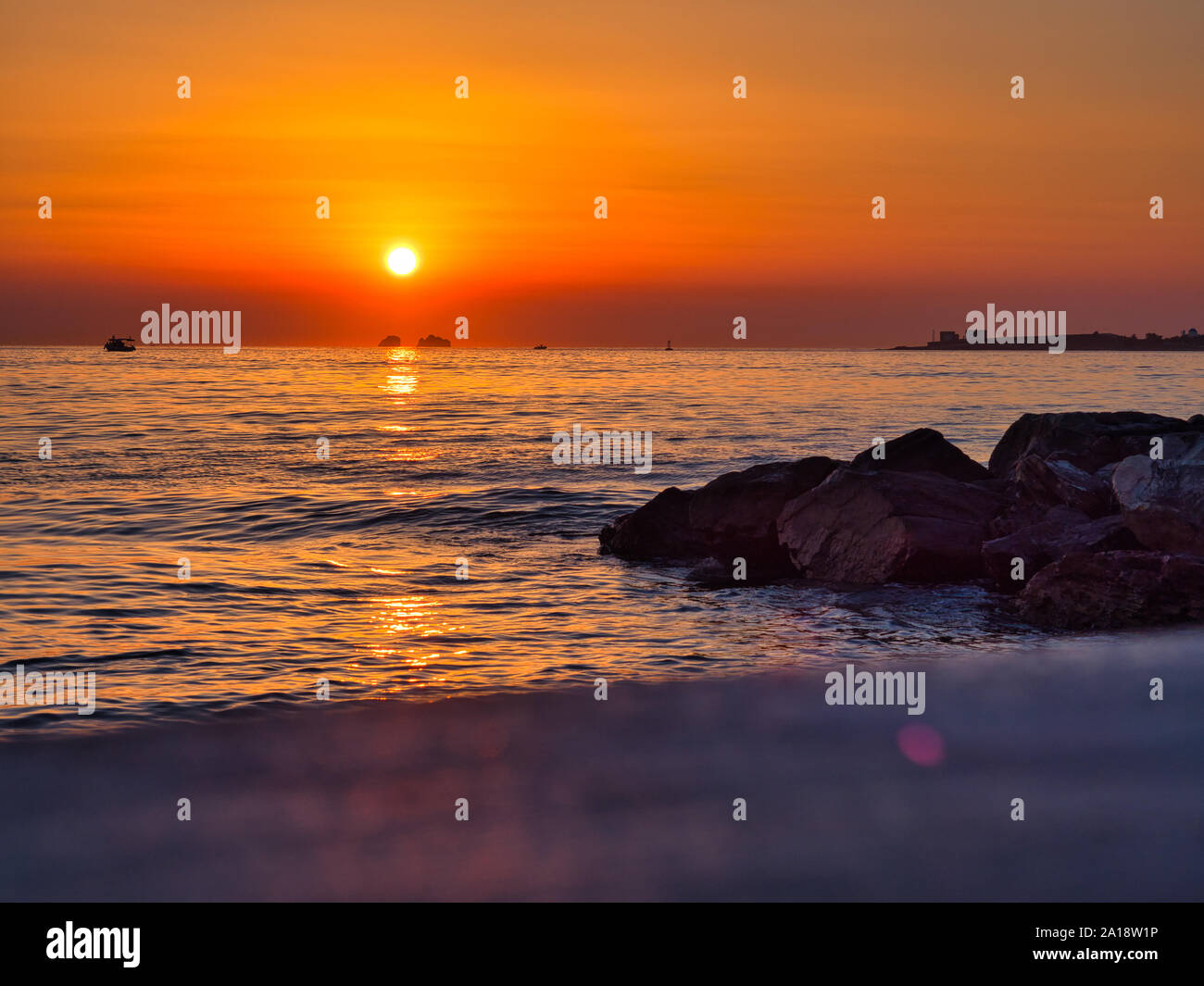 Malerische Landschaft von Orange Sun in bunten Himmel Einstellung hinter dem Wasser mit Booten und Felsen in Parikia Paros Griechenland in Abend Stockfoto