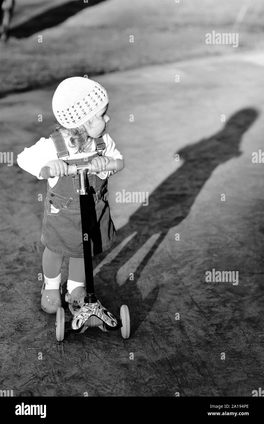 Schwarz-weiß-Porträt einer stilvollen ein Jahr altes Mädchen reiten ein Roller und ihr Schatten an einem schönen Sommertag im Park Stockfoto
