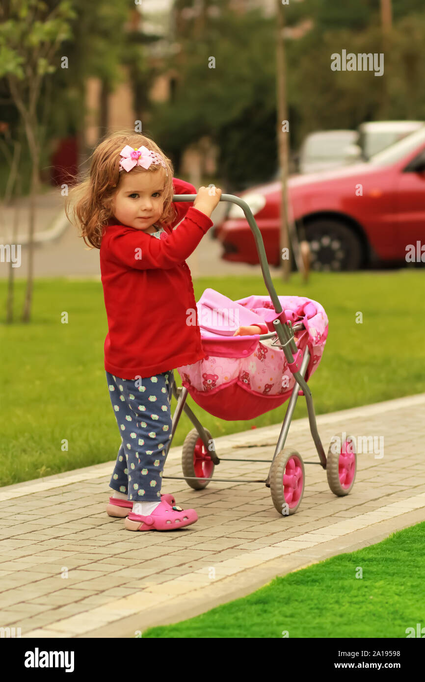 Porträt eines ein Jahr alten niedlichen kleinen curly Girl das Tragen der roten Strickjacke und ein Kopfband mit einem Bogen mit einem Spielzeug rosa im Park ihr Motte emulieren Spaziergang Stockfoto