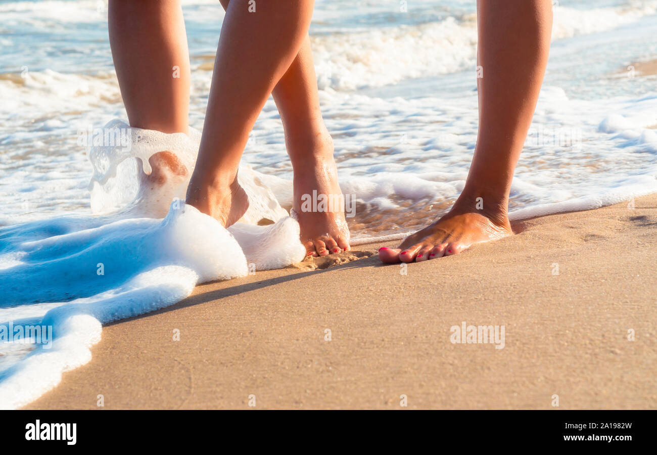 Weibliche Füße an einem Sandstrand in die Wellen der Brandung des Meeres im Sommer Nahaufnahme Stockfoto