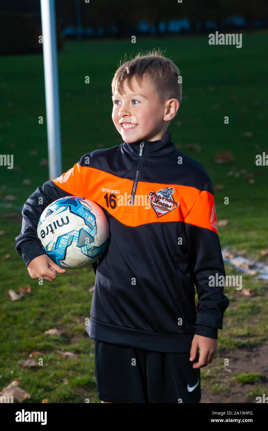 Ein acht Jahre alter Junge tragen ein Fußball-Streifen und halten ein Fußball unter dem Arm. Stockfoto