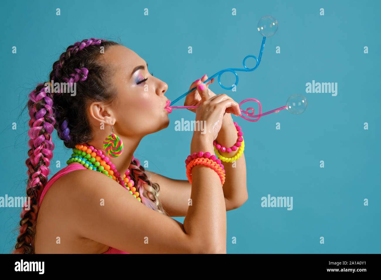 Schöne Mädchen mit einem bunten Zöpfe Frisur und Make-up, bläst Seifenblasen mit Tubuli, im Studio vor einem blauen Hintergrund posiert. Stockfoto