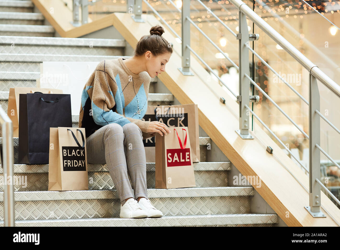 Junge Frau in Freizeitkleidung sitzt auf der Treppe und Suchen in den Warenkorb legen nach dem Shopping in der Shopping Mall Stockfoto