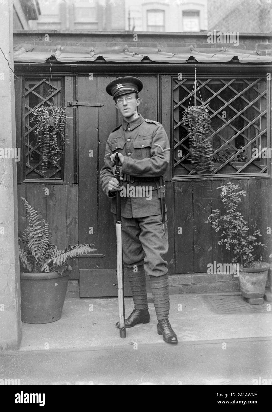 Ein Vintage schwarz-weiß Foto zeigt einen jungen Mann, Tragen einer Brille im Ersten Weltkrieg Soldat der britischen Armee, Uniform gekleidet, im Hinterhof eines Hauses stehen, lehnte sich auf seinem Gewehr. Stockfoto