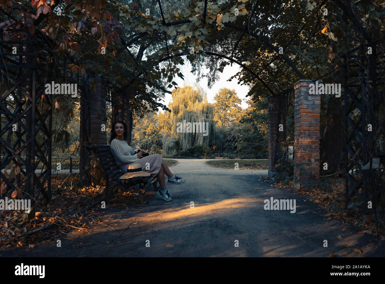 Frau in romantischen Kleidung und Strohhut im Freien im Herbst Park und genießen Sie gutes Wetter und Sonnenuntergang, beim Sitzen auf der Bank unter gotischen Bogen Stockfoto