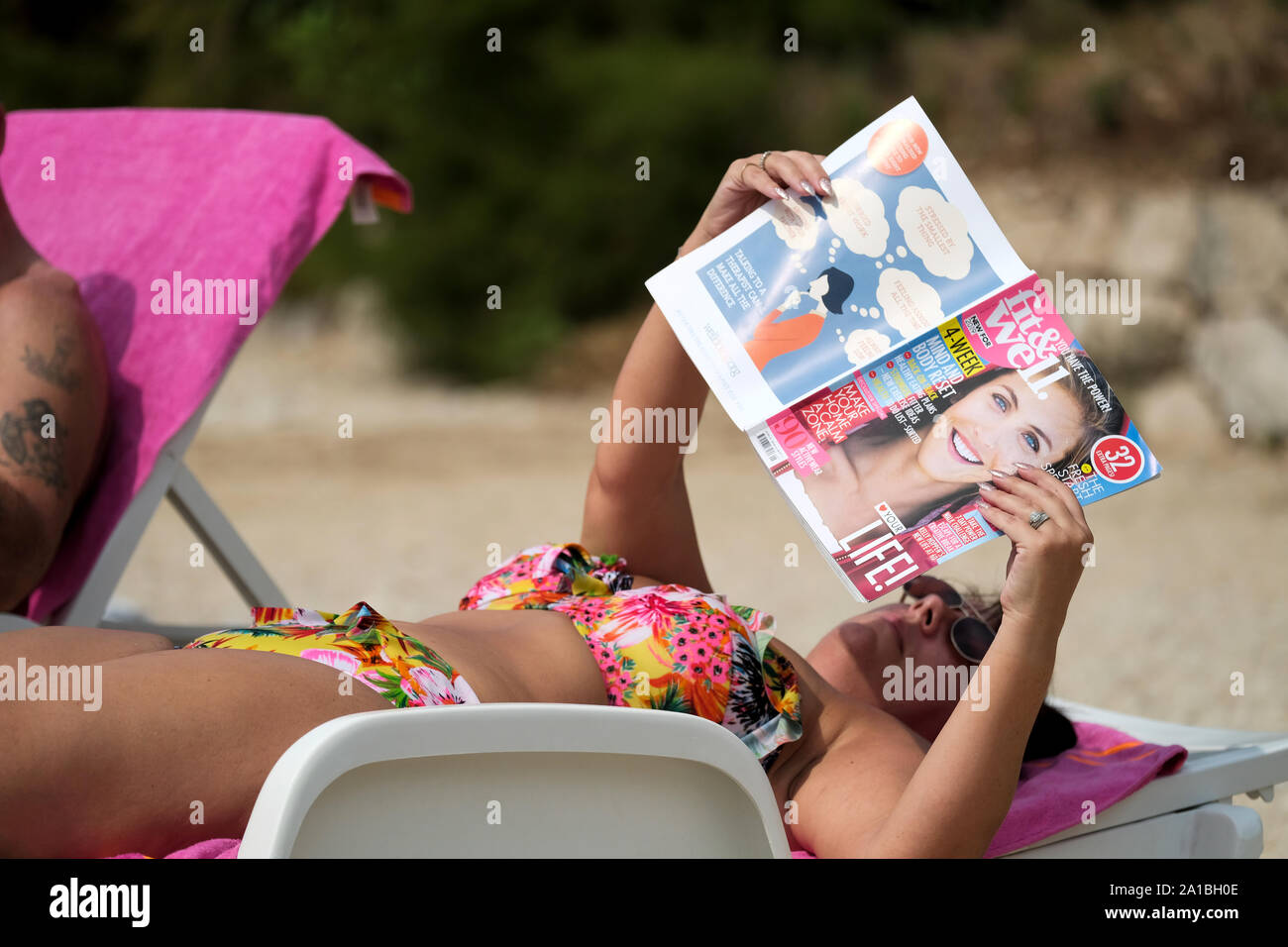 Eine Frau mittleren Alters in der Sonne entspannen, am Strand und das Tragen eines Bikini. Sie liest eine beliebte englische Magazin Fit und Gut Stockfoto