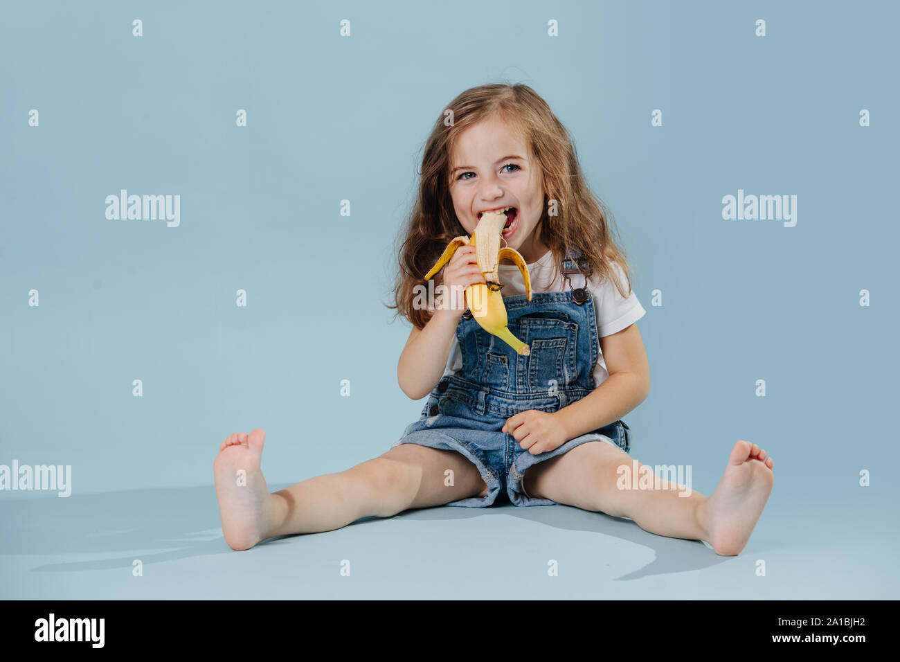 Kleine Mädchen essen Bananen, während auf dem Boden über blauen Hintergrund sitzen Stockfoto
