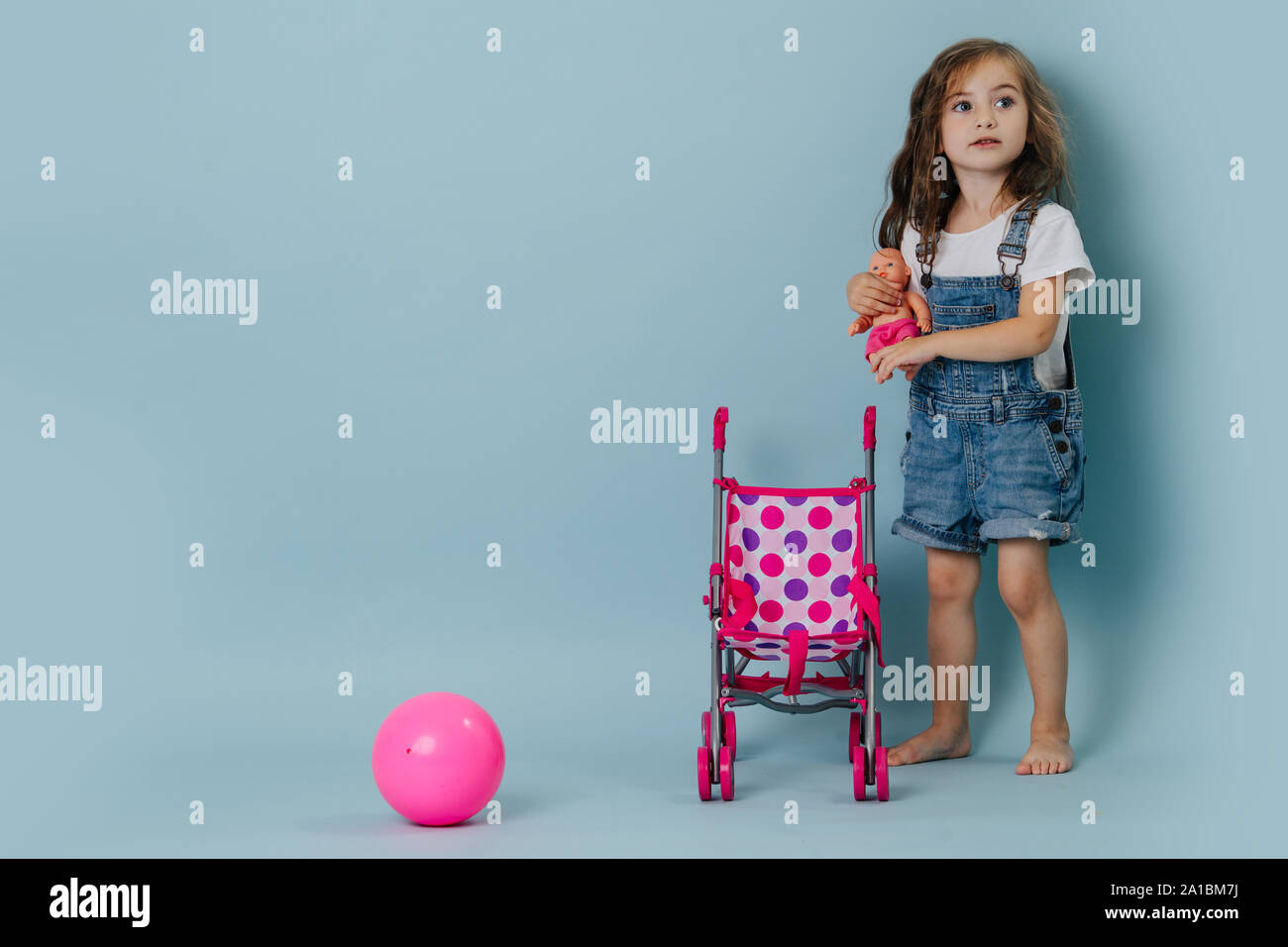 Kleines Mädchen eine Puppe hält neben einem Kinderwagen und rosa Gymnastikball über Blau Stockfoto
