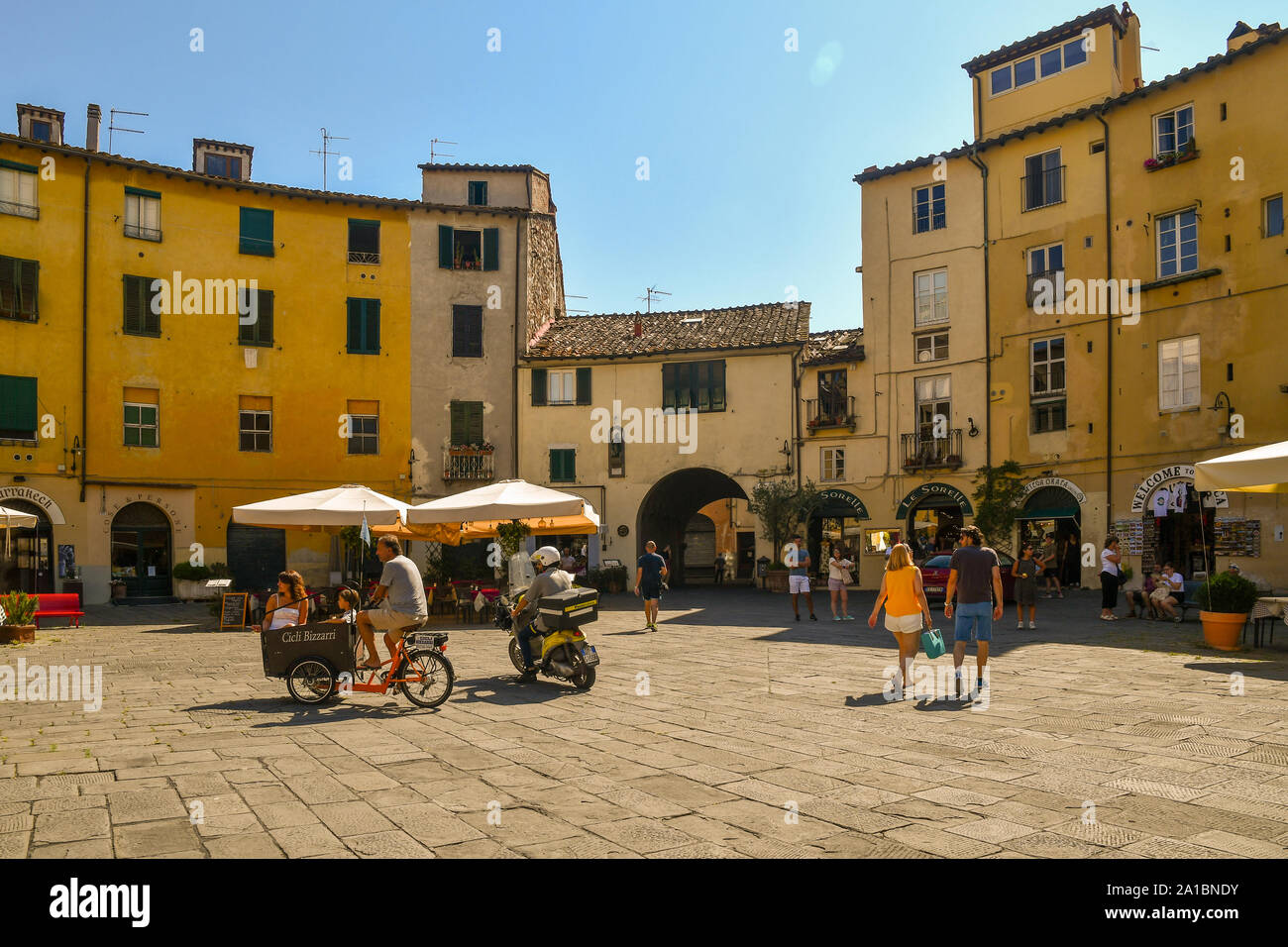 Piazza dell'Anfiteatro, einem berühmten elliptische Platz in Lucca, mit Touristen, eine Familie auf einer cargo bike und ein Briefträger auf einem Roller, Toskana, Italien Stockfoto