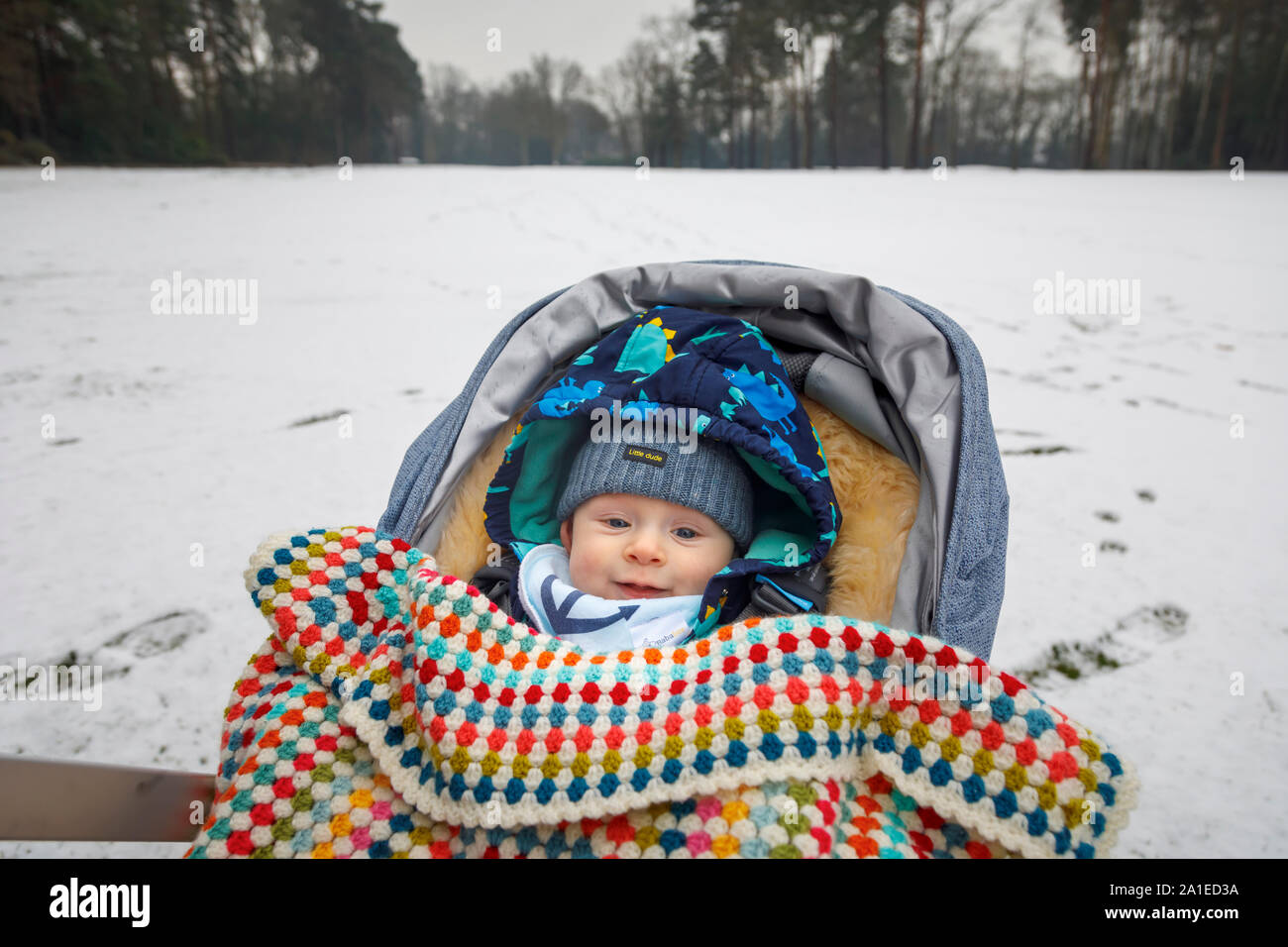 Ein kaukasischer Baby zwischen 4 und 5 Monate alt, mit blauen Augen im Kinderwagen im Winter durch eine gehäkelte Decke tragen einer Schirmmütze abgedeckt im Alter von Stockfoto