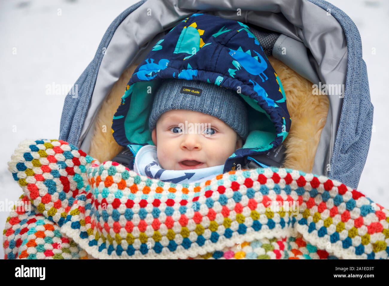 Ein kaukasischer Baby zwischen 4 und 5 Monate alt, mit blauen Augen im Kinderwagen im Winter durch eine gehäkelte Decke tragen einer Schirmmütze abgedeckt im Alter von Stockfoto