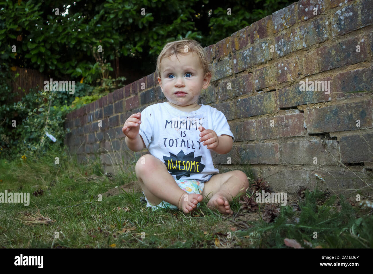 Ein schlammiges kleinen kaukasischen Jungen (Alter 11 Monate) mit blauen Augen das Tragen eines grubby weißes T-Shirt oben sitzt im Garten, der von einer Ziegelmauer besorgt suchen Stockfoto