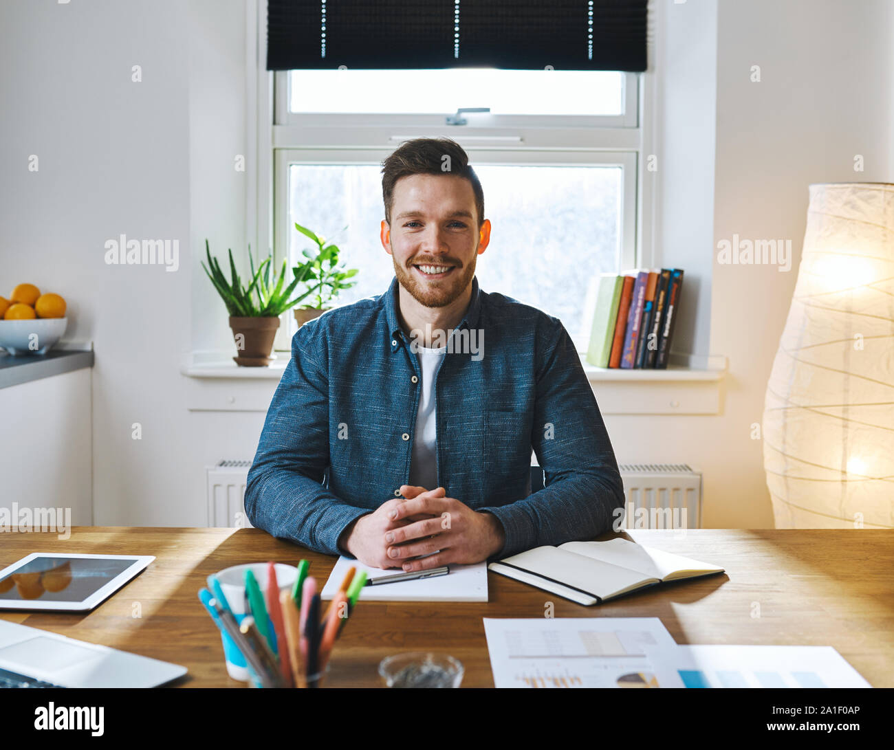 Schöner Lächeln bärtiger Mann mit gefalteten Händen in legere Kleidung am Schreibtisch sitzen vor dem hellen Fenster Stockfoto