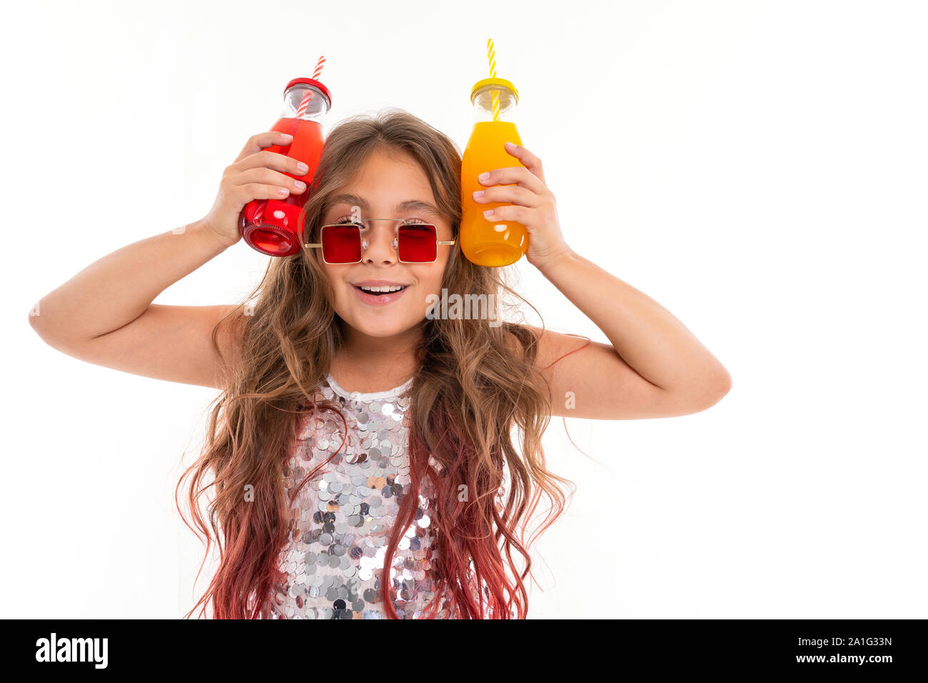 Happy girl im glitzernden Kleid und stilvolle Quadratische rote Sonnenbrille Herumfummeln zwei Glasflaschen: Mit roten trinken und mit gelben Drink isoliert Stockfoto