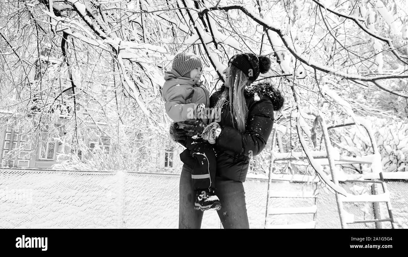 Monohrome Porträt der Mutter mit ihrem süßen Sohn in Jacke und Hut spielen mit Schnee bedeckten Baum auf dem Spielplatz im Park Stockfoto