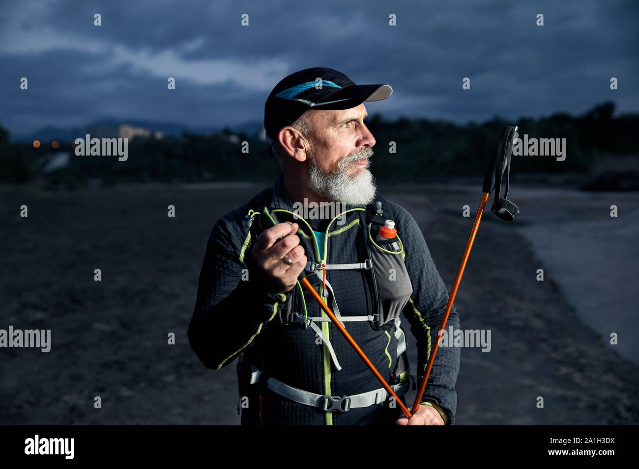 Portrait von älteren Athleten Mann mit grauem Bart und Stöcke an dunklen bewölkter Himmel Hintergrund Stockfoto