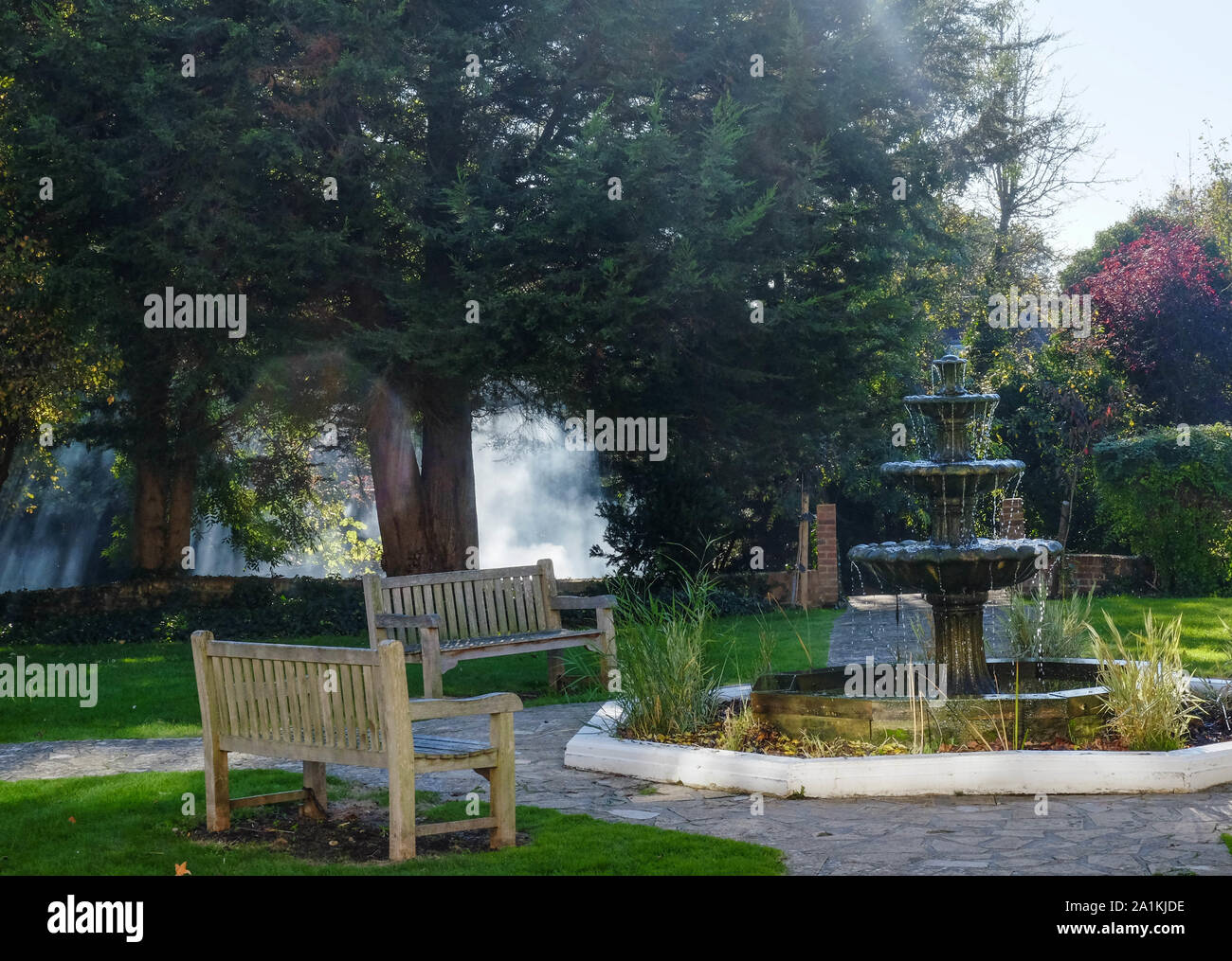 Ruhigen Garten im Herbst mit Holzbänken, 3 Stufe Stein Wasser Brunnen, grünes Gras und Bäume im Hintergrund. Stockfoto