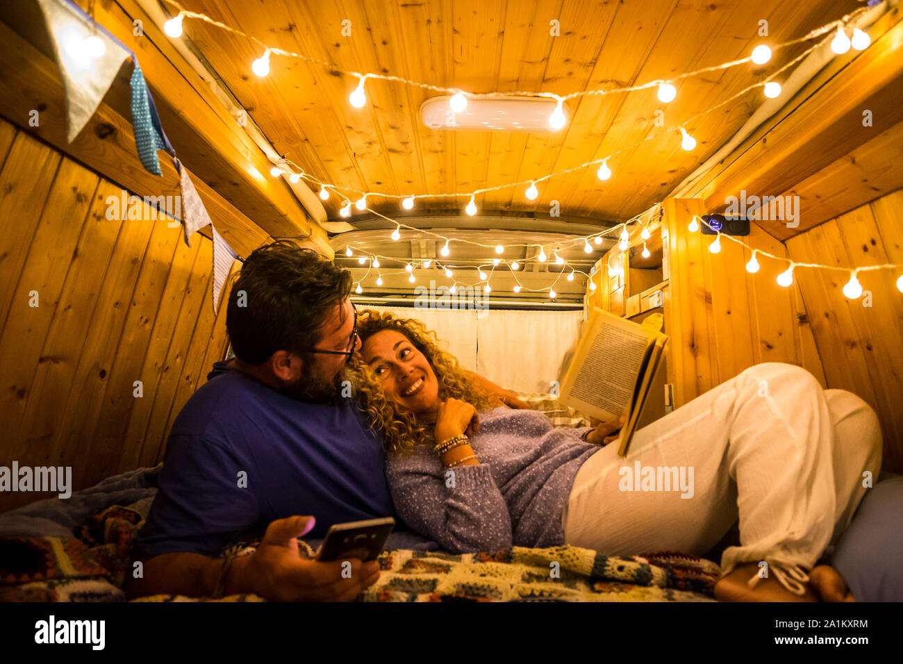 Paar Erwachsene leben in einer schönen und Recht van unten auf dem Bett liegen, ein Buch lesen und mit niedlichen leuchtet auf ihre Köpfe - vanlife l Stockfoto