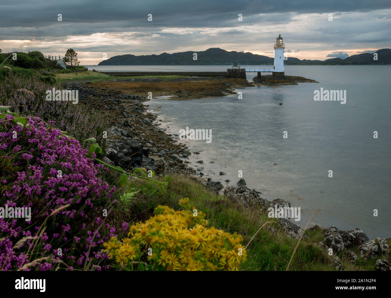 Ausblicke auf die Landschaft. Tobermory Leuchtturm bei Sonnenuntergang. Isle of Mull. Moody bewölkter Himmel über den Bergen im Hintergrund. Gelb und Violett wilden Blumen in foregr Stockfoto
