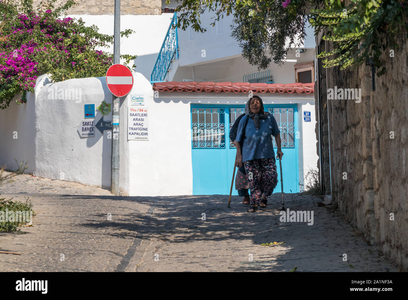 Alacati, Türkei - 4. September 2019: Alte Frau mit Stöcken wandern in einer engen Straße. Die Stadt ist bei Touristen beliebt im Sommer Stockfoto