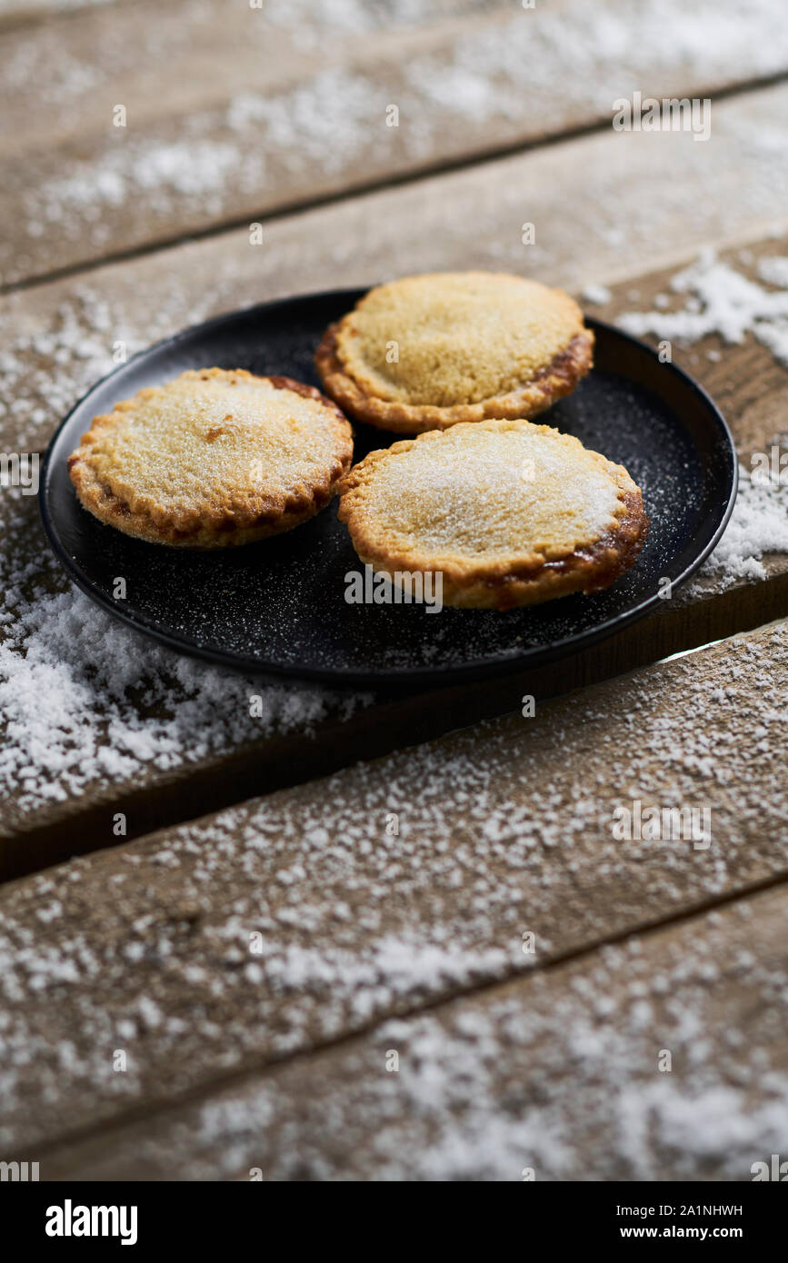Drei Mince Pies auf einer schwarzen Platte auf einem rustikalen Holztisch mit Schnee Stockfoto