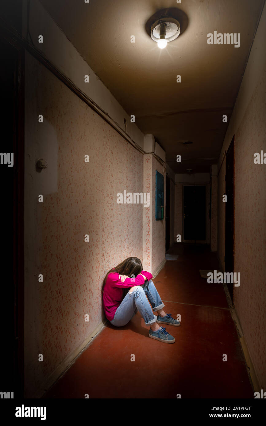 Eine traurige und verzweifelte Frau sitzt in einem dunklen Korridor durch ein düsteres Licht beleuchtet. Ihr Schmerz und ihre vielen Probleme drückte sie in die völlige Isolation. Hi Stockfoto