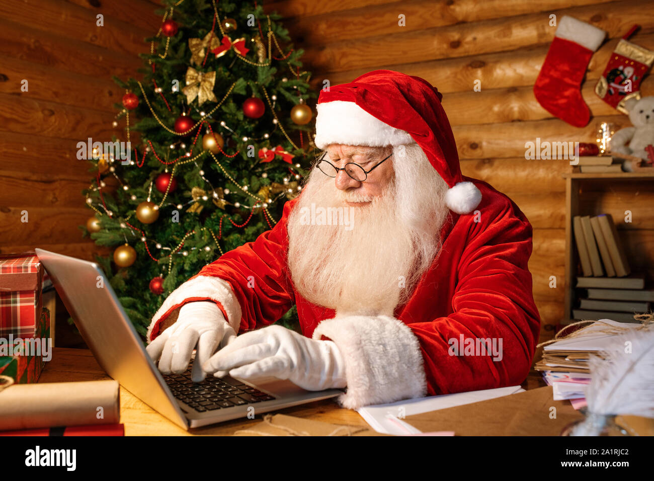 Zeitgenössische Santa Tippen auf Laptop keypas während der Sitzung von Tisch und auf der Suche nach online Shops für Weihnachtsgeschenke kaufen Stockfoto