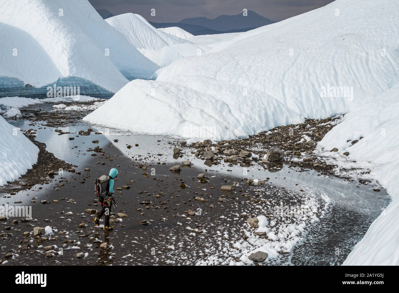 Tief im Hinterland in Alaska auf der Matanuska Gletscher, eine junge, weibliche Eiskletterer fließt durch einen breiten, felsigen Tal voller seichtem Wasser. Stockfoto