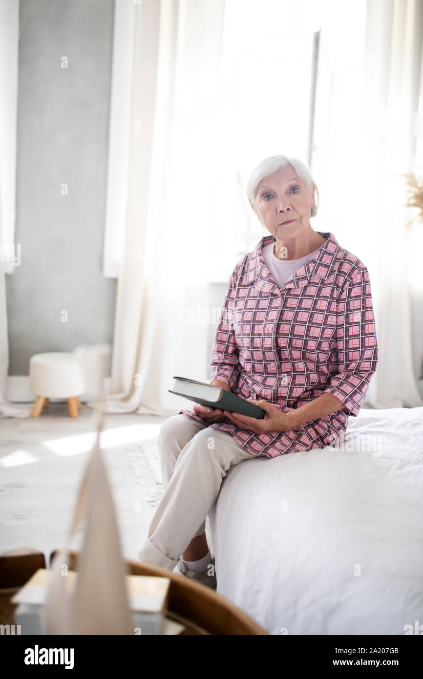 Grauhaarige Frau sitzt im Schlafzimmer mit Buch Stockfoto