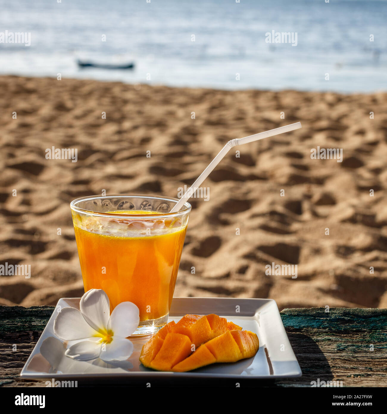 Mango Saft serviert mit einer Hälfte der Mango und eine Frangipani Blume auf den Strand und das Meer im Hintergrund. Indonesien. Quadratisches Bild. Stockfoto