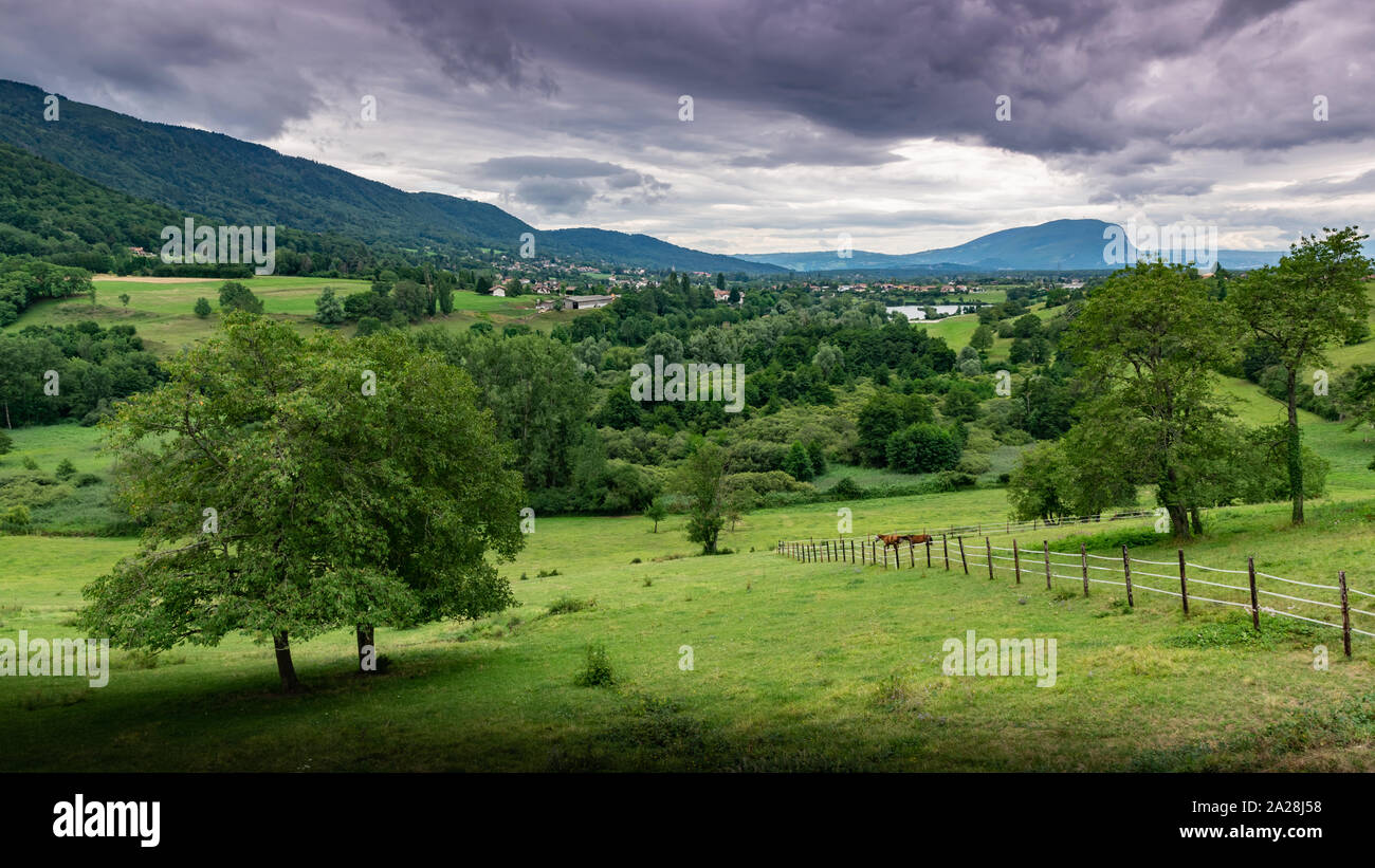 Landschaft der Berge, Wälder, kleine Stadt, grüne Wiesen und Zwei Pferde auf einer eingezäunten Weide auf dem Hintergrund eines bewölkten Himmel. Machilly. Stockfoto