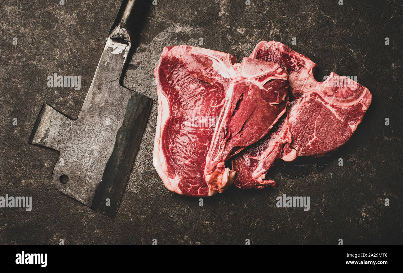 Flachbild-lay von frischem nicht gegartem Rindfleisch T-Bone Steaks und Metall Metzger cleaver über rauhe schwarze Stein Hintergrund, Ansicht von oben. Metzger Shop Konzept Stockfoto