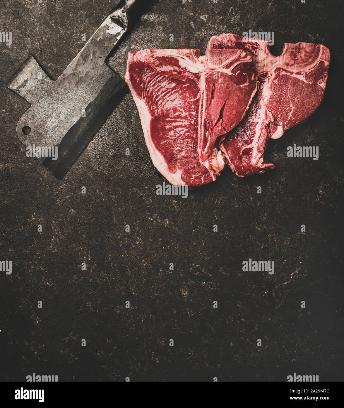 Flachbild-lay von frischem nicht gegartem Rindfleisch T-Bone Steaks und Metzger cleaver über rauhe schwarze Stein Hintergrund, Ansicht von oben, kopieren. Metzger Shop Konzept Stockfoto