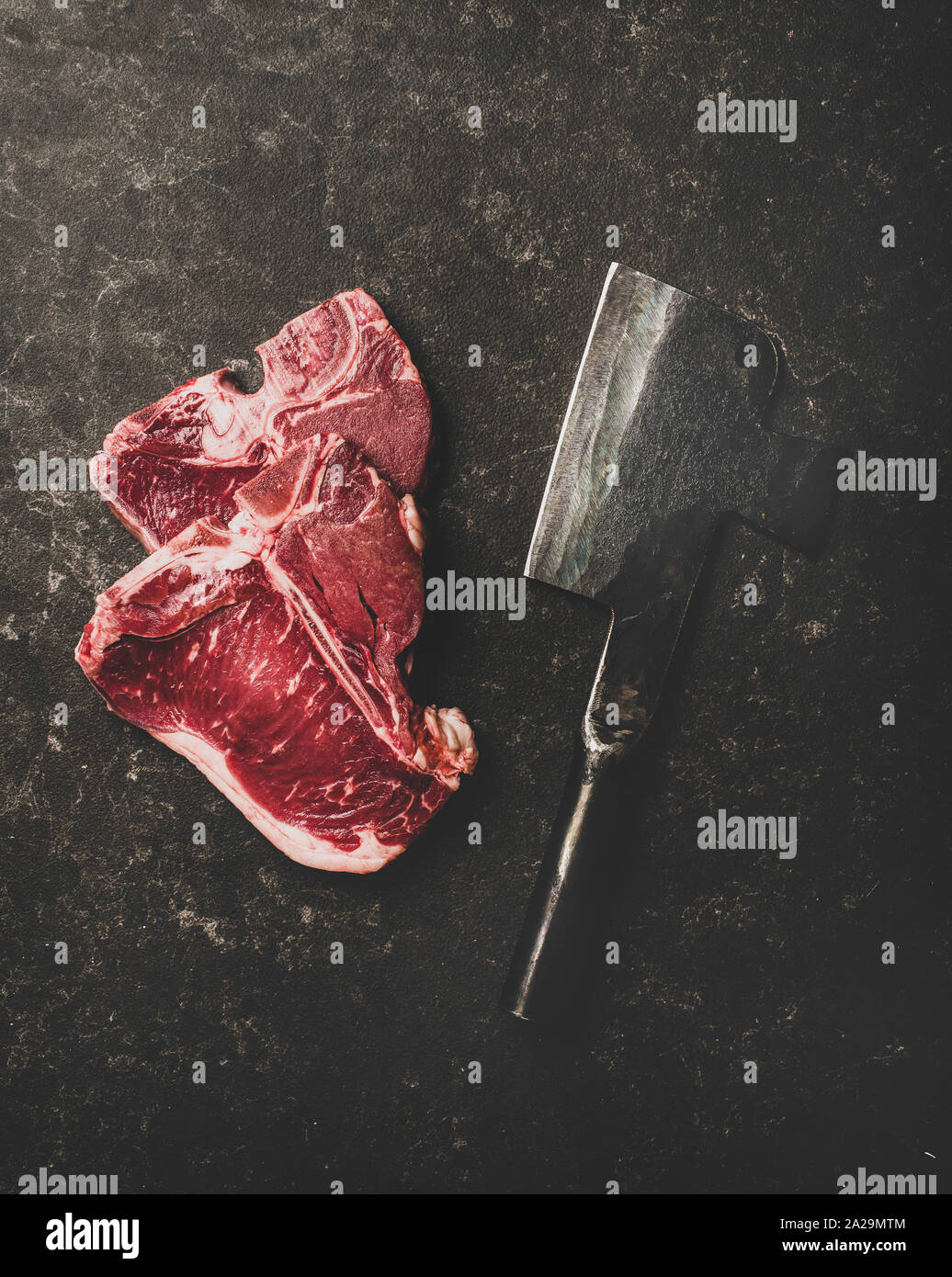 Flachbild-lay von frischem nicht gegartem Rindfleisch T-Bone Steaks und Metzger cleaver über rauhe schwarze Stein Hintergrund, Ansicht von oben. Metzger Shop Konzept Stockfoto