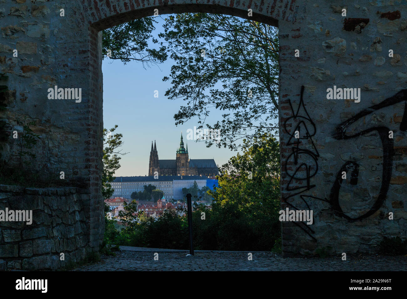 Die Prager Burg aus der Sicht Petrin im Stadtteil Mala Strana an einem sonnigen Tag mit blauem Himmel mit Blick durch einen steinernen Tor und Bäume und Sträucher Stockfoto