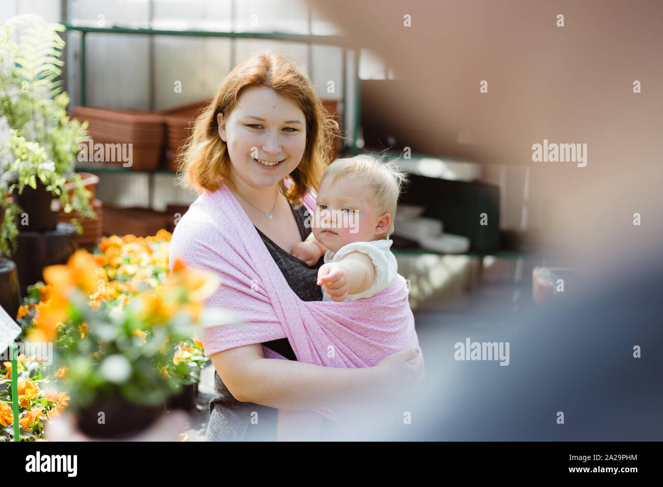 Nahaufnahme der Mutter, die ihr Baby Tochter in einem rosa Sling wickeln Sie beim Einkauf für Blumen für Ihren Garten in einem lokalen Gewächshaus Stockfoto