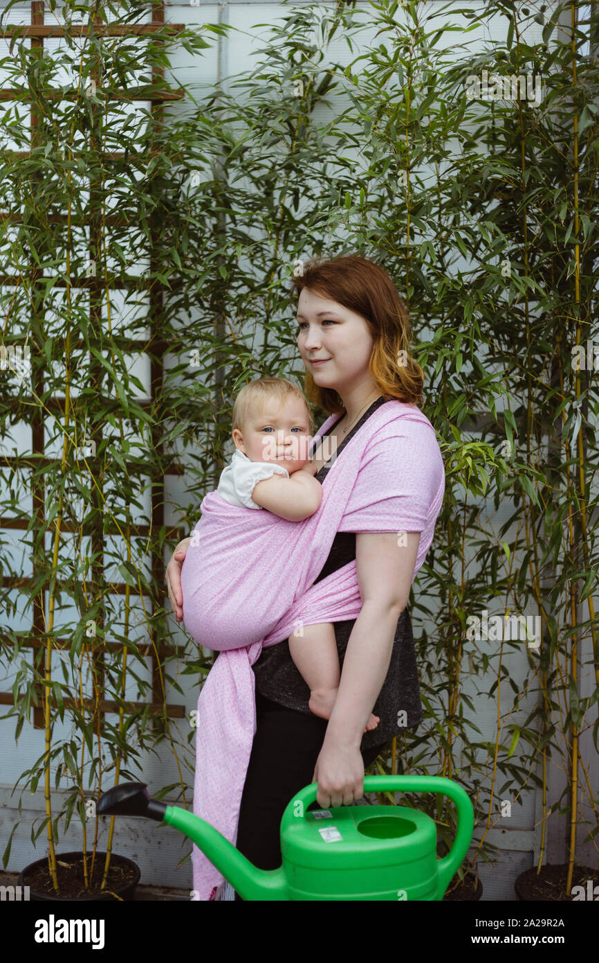 Junge rothaarige babywearing Mutter tragen sie verschlafen ein Jahr alten Baby Mädchen in rosa wrap Schlinge bei der Gartenarbeit Stockfoto