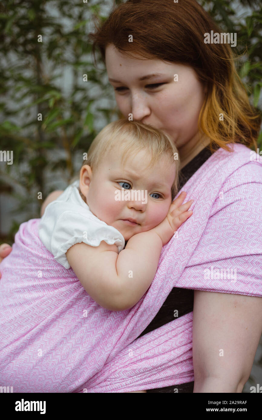 Junge rothaarige babywearing Mutter tragen sie verschlafen ein Jahr alten Baby Mädchen in rosa wrap Schlinge Stockfoto