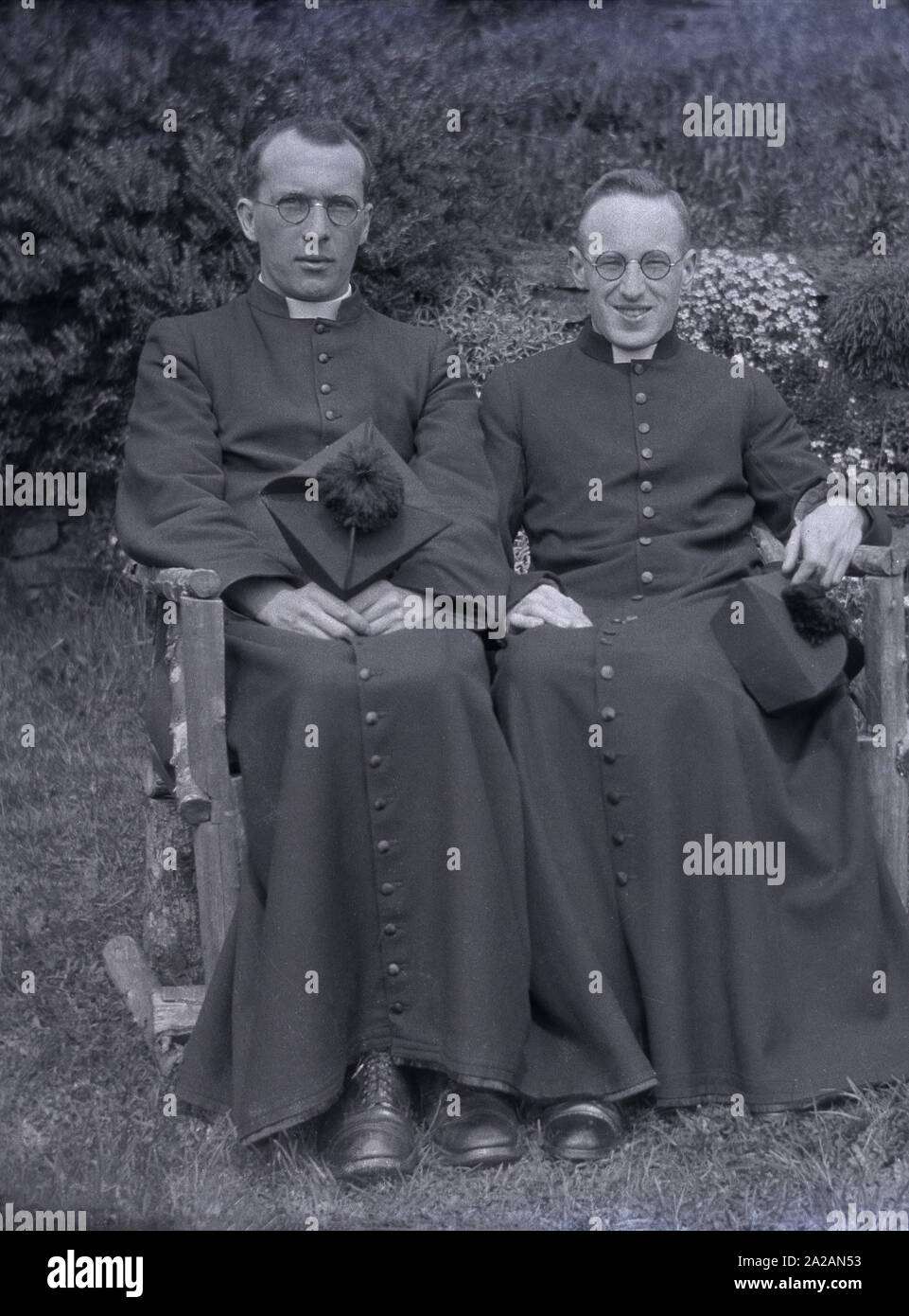 Um die 1920er Jahre, historisch, zwei römisch-katholische Priester, die draußen in ihren langen Gewändern auf einer kleinen Bank sitzen, ihre kirchlichen Hüte, eine Biretta, ein steifes quadratisches cape mit vier Graten über der Spitze, überragt von einem Büschel, England, Großbritannien. Die knöchellangen Kleider mit langen Knöpfen werden als Cassocken bezeichnet und sind ein traditioneller christlicher Kirchenmantel. Stockfoto