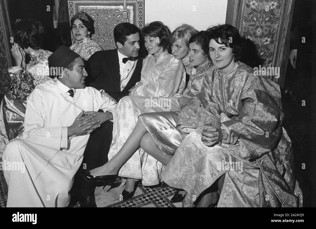 Die Gäste sitzen auf Teppichen und Kissen. Die Männer tragen sowohl europäische als formale Abnutzung und traditionelle arabische Gewänder. Die Damen kombinieren orientalische Kleidung mit Europäischen Schuhe und Frisuren. Stockfoto