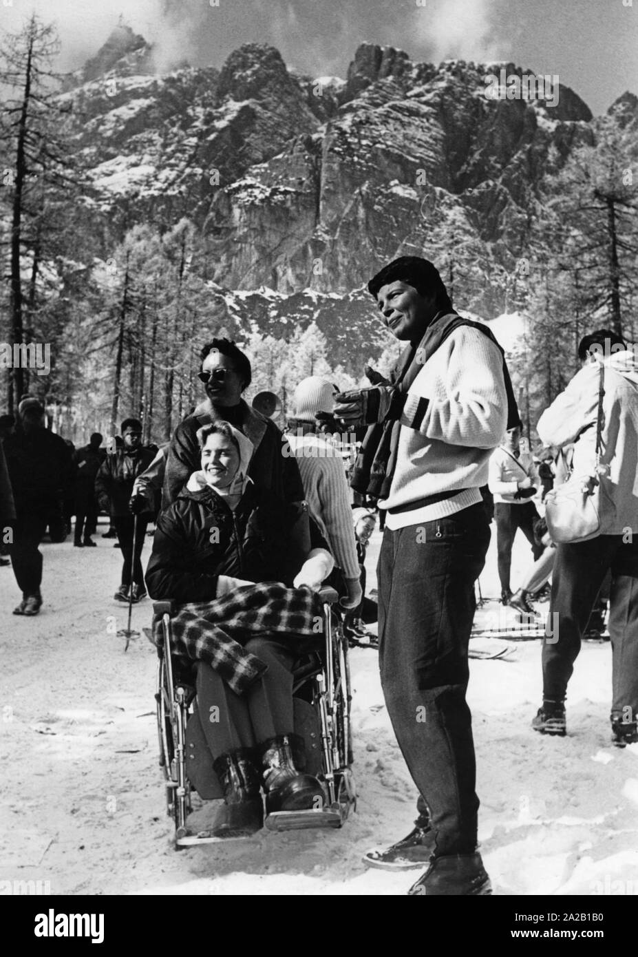 Das Bild zeigt die Besucher der Olympischen Winterspiele 1956 in Cortina d'Ampezzo. Eine Dame in einem Rollstuhl und ihren beiden Gefährtinnen steht im Schnee zwischen den anderen Fans, Sportler, und drücken Sie die Menschen. Stockfoto