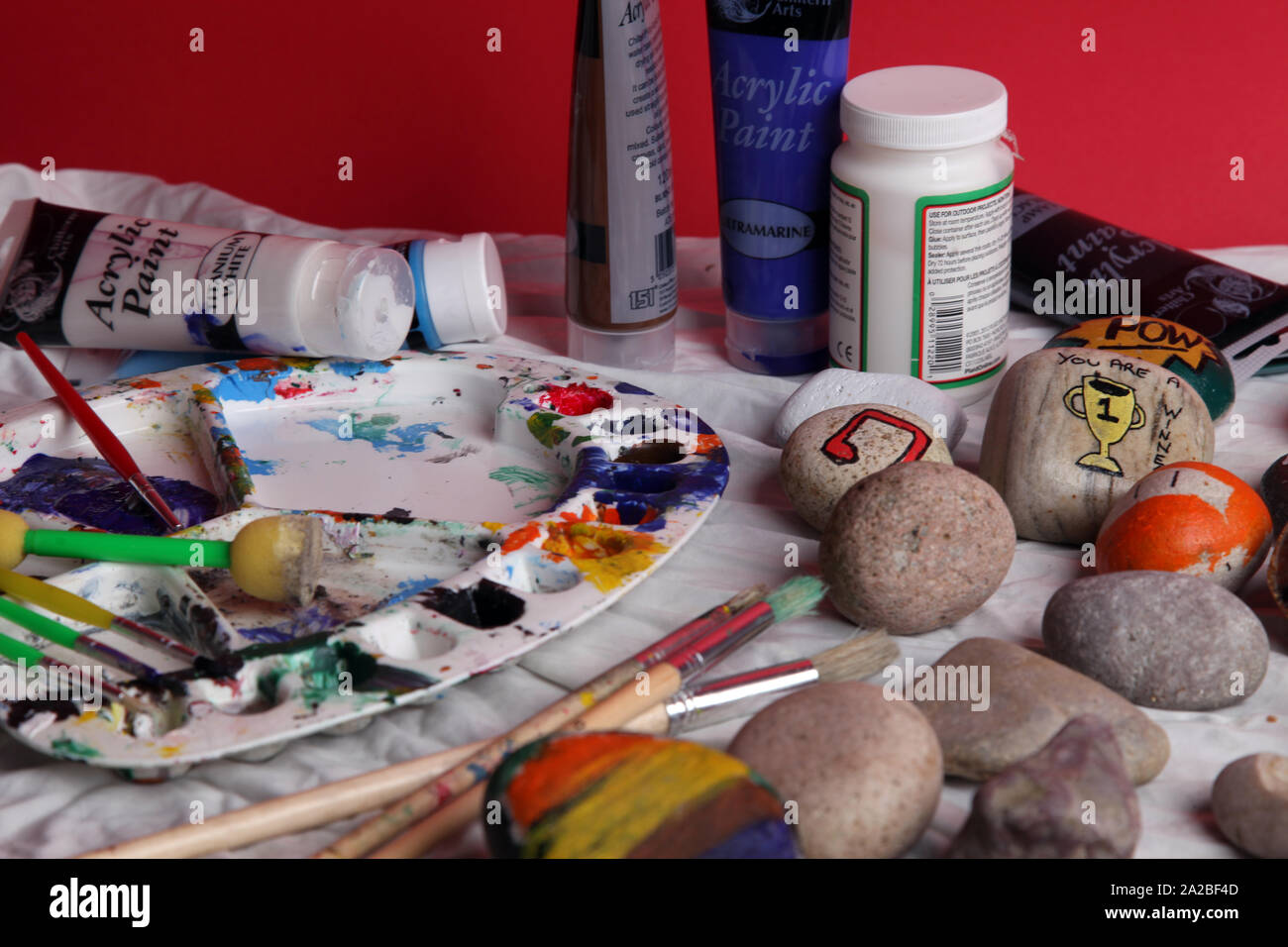 Eine Auswahl von bemalten Felsen auf einer Kunst und Handwerk Tabelle mit Bürsten setup für Rock Malerei als Teil der Rockfun Phänomene auf rotem Hintergrund Stockfoto