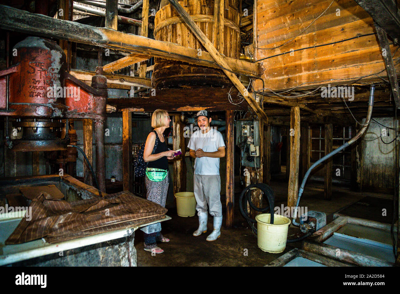 Shohei Nozue erklärt der Reporterin Angela Berg, dass die Moromi im Sommer alle sieben Tage und im Winter alle 14 Tage mit einem großen Paddel gerührt wird. Fermentation und Zeit sind die wichtigsten Komponenten für eine hochwertige Sojasauce. Herstellung von japanischer Sojasauce in 1872 Mejiiya Shoyu Manufaktur in Hamamatsu, Japan Stockfoto