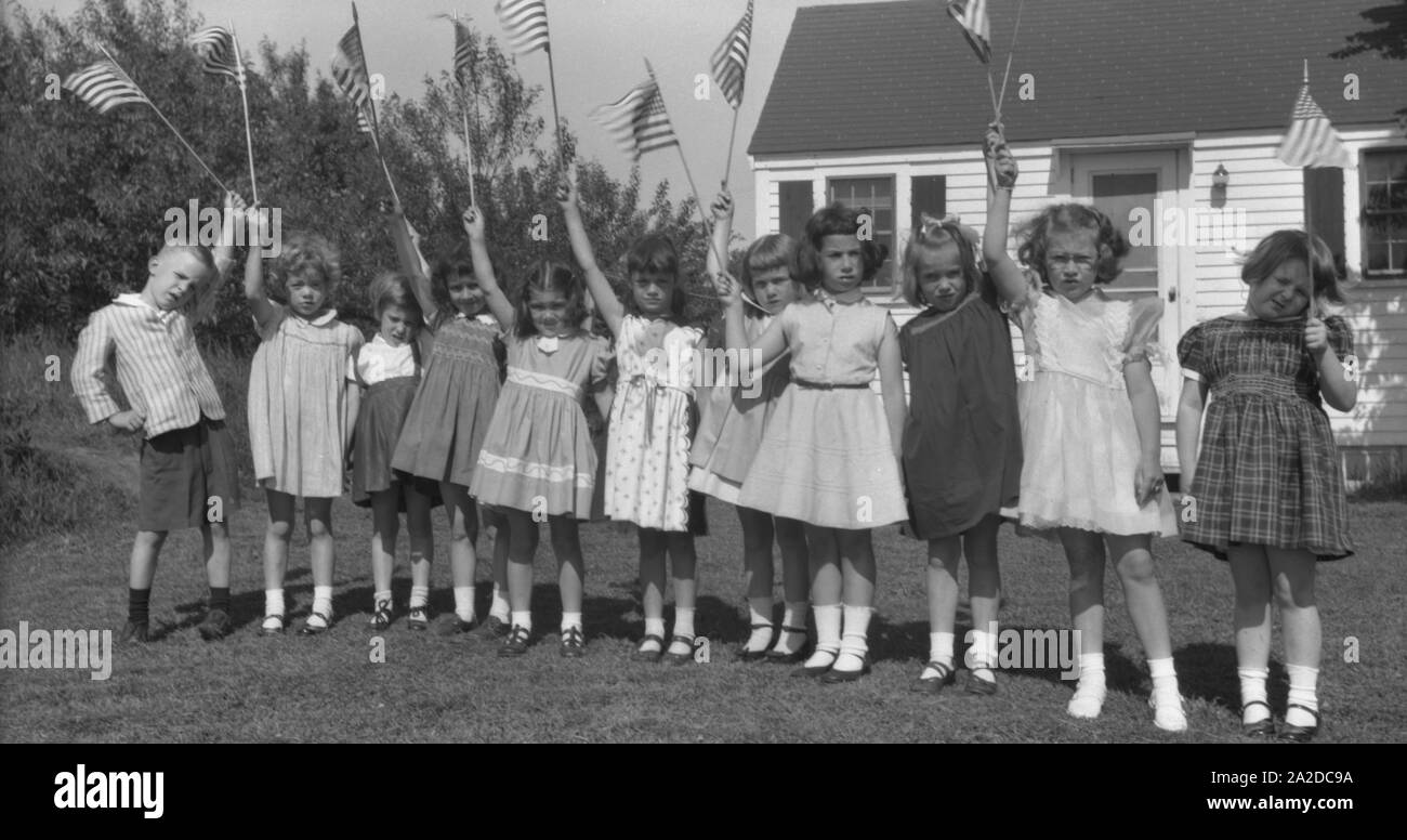 Die 1950er Jahre, die Geschichte, der Sommer und eine Gruppe kleiner Kinder, die auf Gras vor einem hölzernen Bretthaus stehen und amerikanische Fahnen halten, USA. Stockfoto
