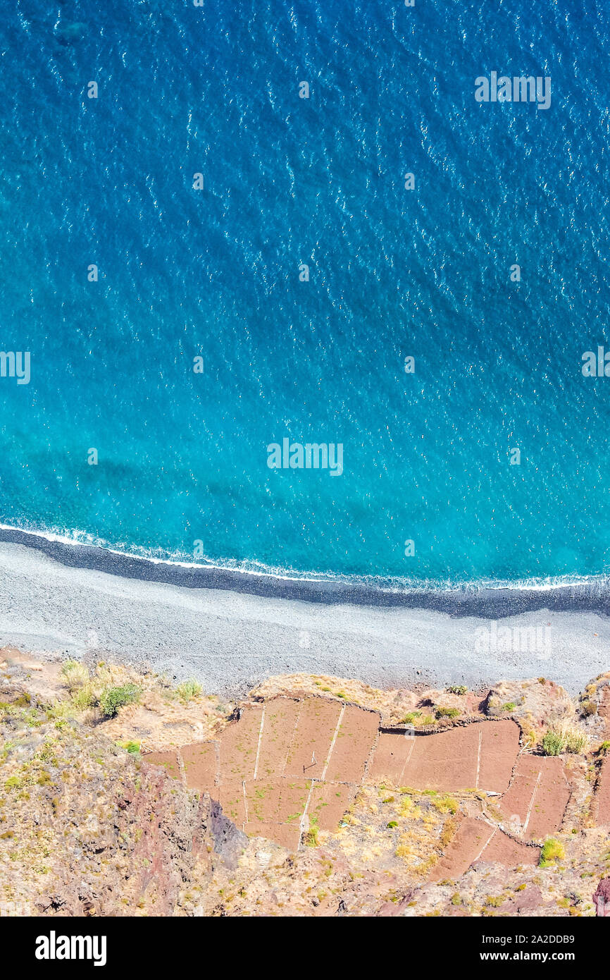 Antenne Landschaft durch den blauen Atlantik. Stein Strand und die angrenzenden Felder an der Südküste der Insel Madeira, Portugal. Luftaufnahme, Meisterwerke der Natur. Sommer Vibes auf einem vertikalen Foto. Stockfoto