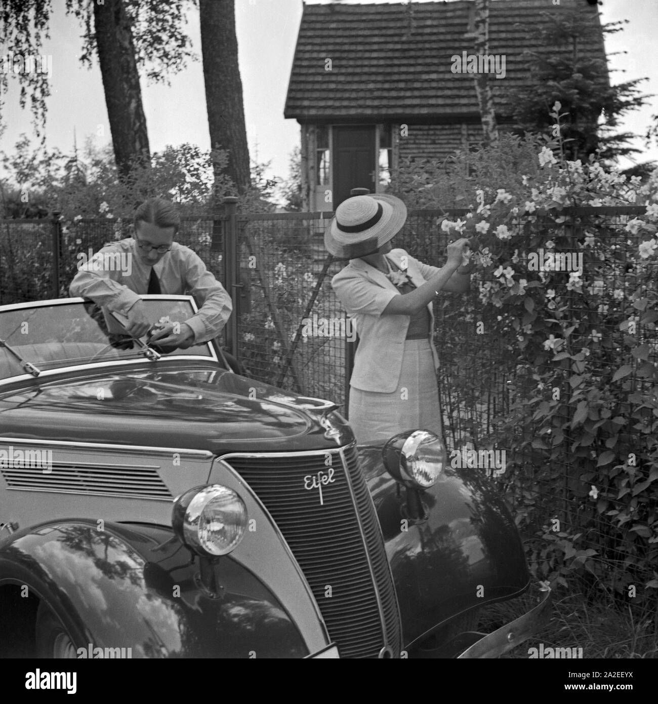 Werbefoto für den Personenkraftwagen Ford Eifel mit einem Paar: der Mann kümmert sich um das Auto, die Frau schaut / Blumen, Deutschland 1935. Kommerzielle Foto für den Pkw Ford Eifel mit ein Paar: der Mann am Auto sieht, die Frau sieht für Blumen, Deutschland 1935. Stockfoto